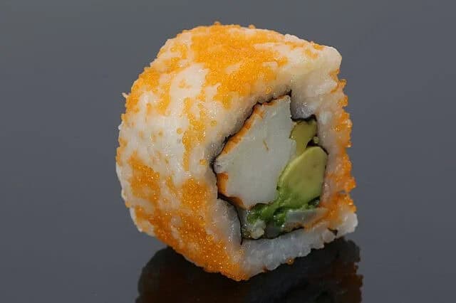 A close-up of a sushi roll topped with bright orange fish roe, filled with avocado, crab stick, and cucumber, set against a dark background.